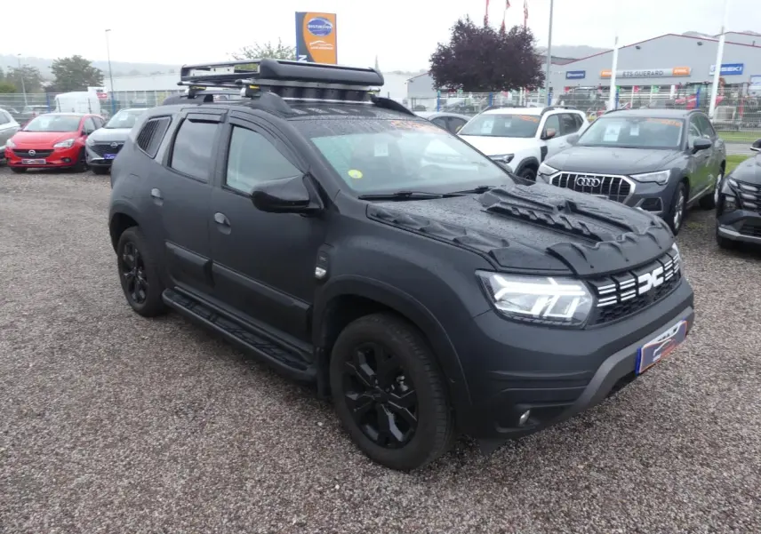 Dacia Duster noir 2023 en 3/4 avant droit avec galerie de toit, marchepieds et jantes noires sur parking.