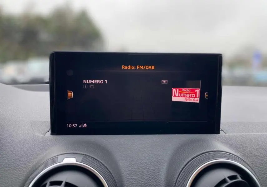 Écran central de l'Audi Q2 2023 affichant la radio FM/DAB, avec deux aérateurs ronds en aluminium visibles en dessous.