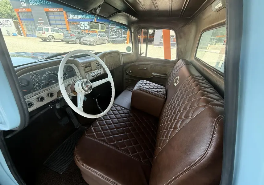 Vue intérieure côté conducteur du Chevrolet C10 Apache 1963 bleu, avec volant blanc et sièges marron matelassés.