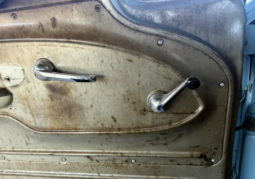 Gros plan sur la poignée de porte intérieure chromée et la manivelle de vitre d’une Chevrolet C10 Apache 1963 bleu, côté gauche.