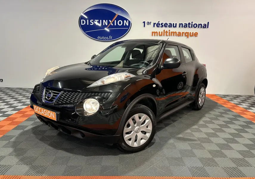 Nissan Juke noir métal en 3/4 avant droit, avec attelage visible, sur sol damier gris et orange en intérieur showroom.