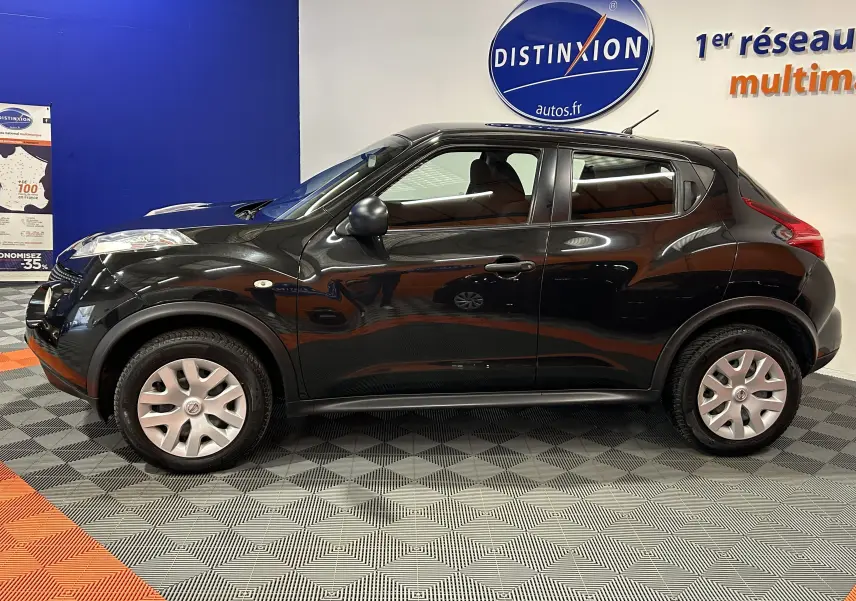 Profil côté gauche d'un Nissan Juke noir métal de 2012 avec attelage visible, posé en intérieur sur sol damier.