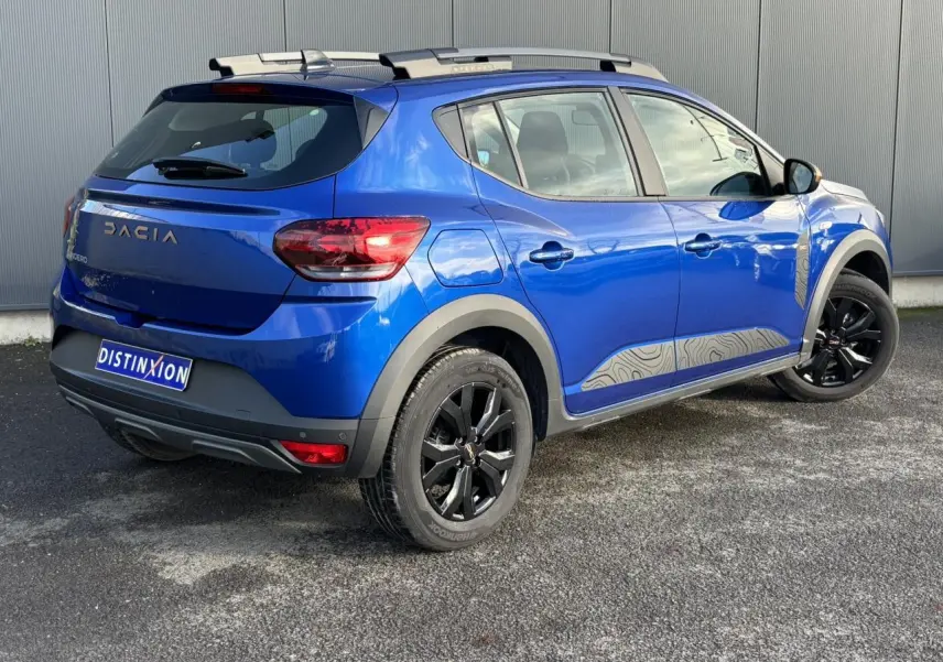 Vue 3/4 arrière droite d'une Dacia Sandero Stepway Bleu Iron avec jantes noires et barres de toit grises.