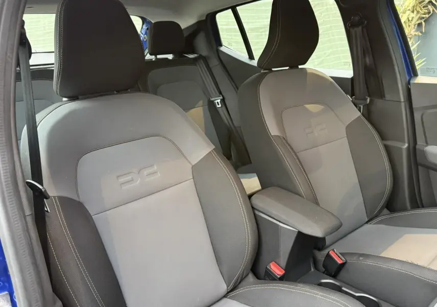 Vue intérieure sur les sièges avant gris avec logo Dacia embossé et accoudoir central, portière bleu visible.