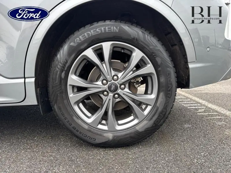 Gros plan sur la roue avant droite en jante alu gris d’un Ford Kuga gris Solar métallisé, avec logo Ford visible au centre.