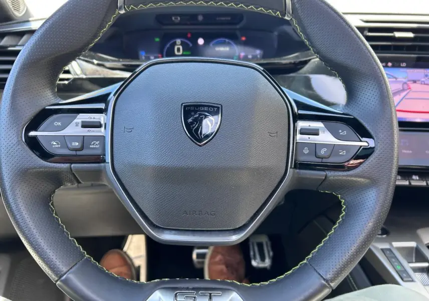 Vue rapprochée du volant cuir noir avec logo Peugeot et badge GT, tableau de bord numérique en arrière-plan.