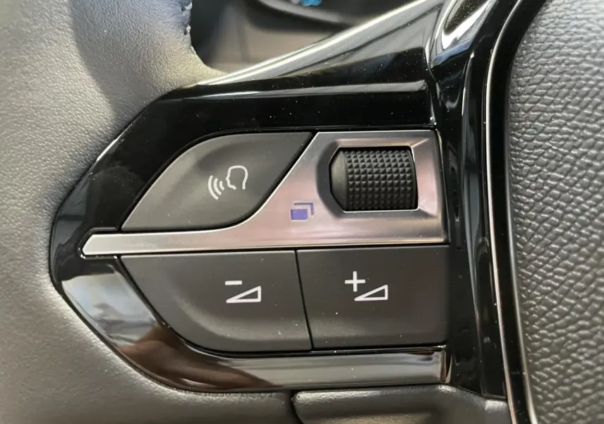 Gros plan sur les commandes de volume et reconnaissance vocale du volant noir du Peugeot 2008 blanc hybride 2026.