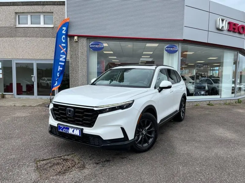 Honda CR-V blanc platine nacré vu en 3/4 avant droit devant un concessionnaire avec toit noir et jantes foncées.