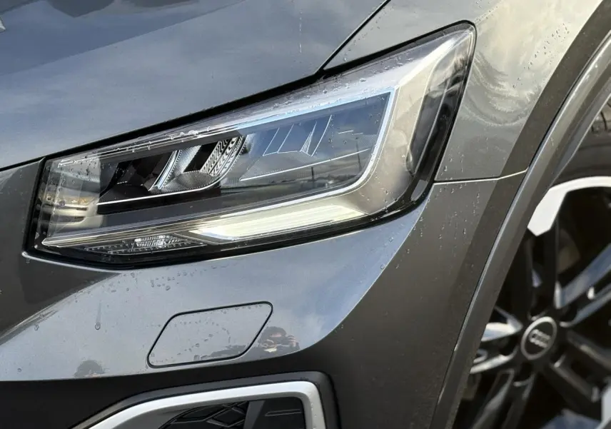 Gros plan sur le phare avant droit et la jante Audi Sport 5 branches titane mat du Q2 gris Daytona 2025.