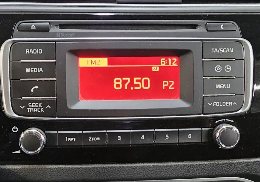 Gros plan sur la radio avec écran rouge affichant la fréquence 87.50 FM dans le tableau de bord noir d'une Kia Rio III 2015.