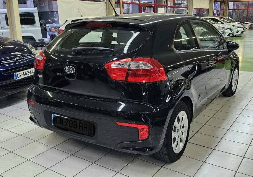 Vue 3/4 arrière droite d'une KIA RIO III noire 3 portes, avec feux arrière rouges et jantes blanches en showroom.