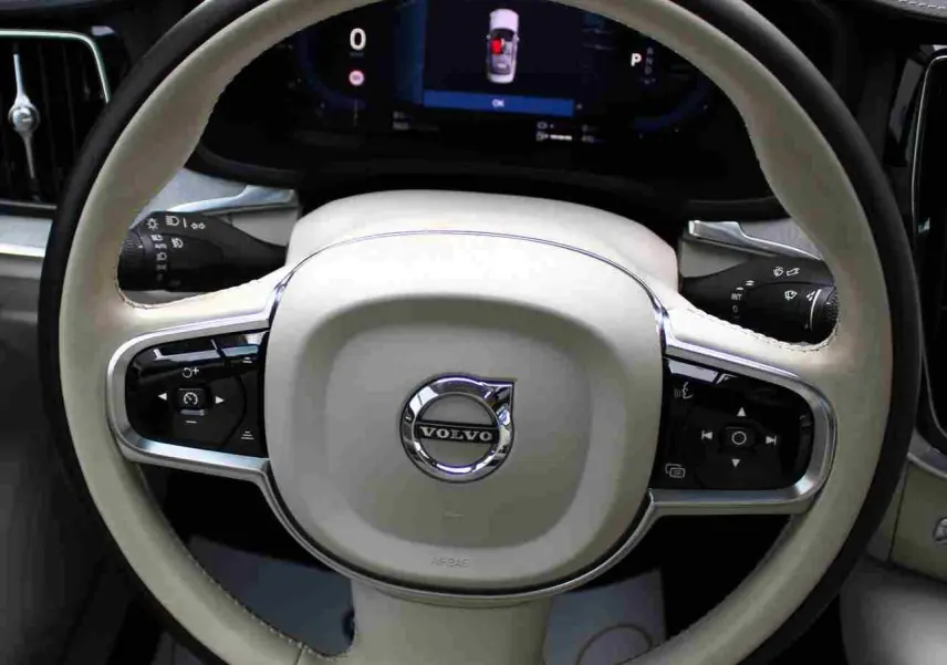 Gros plan sur le volant beige clair de la Volvo S60 2024 hybride rechargeable avec commandes intégrées et tableau de bord numérique.