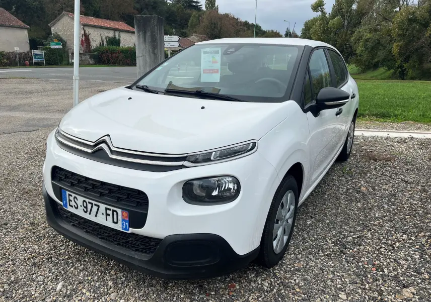 Citroën C3 blanc vue 3/4 avant droit, avec calandre distinctive et phares ronds, garée sur gravier en extérieur.