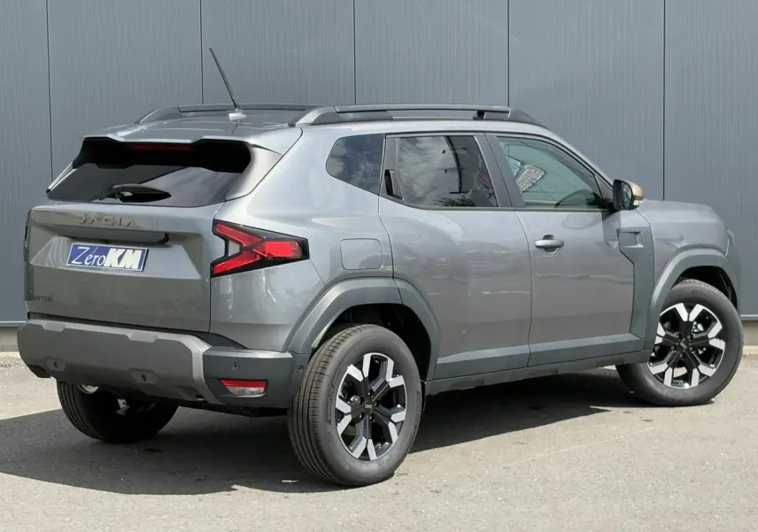 Dacia Duster gris schiste en 3/4 arrière droit, avec jantes alliage noires et coques de rétroviseur brun cuivré.