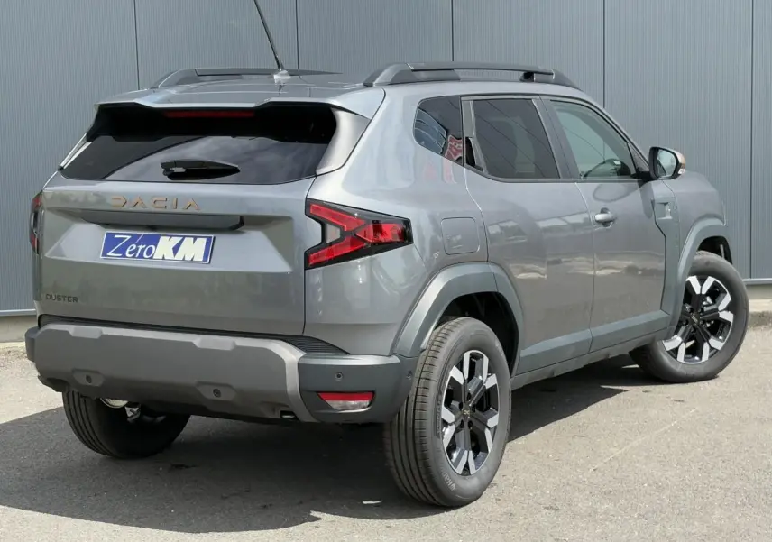 Vue 3/4 arrière droite du Dacia Duster 2025 gris schiste avec jantes alliage et barres de toit noires.