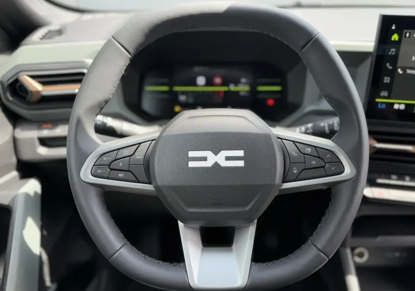 Volant multifonctions noir et gris du Dacia Duster 2025 avec tableau de bord digital et écran tactile visible en arrière-plan.