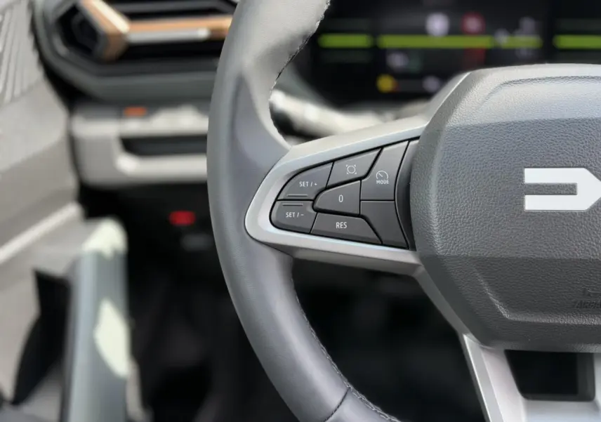 Gros plan sur le volant gris du Dacia Duster 2025, avec commandes intégrées et tableau de bord digital flou en arrière-plan.