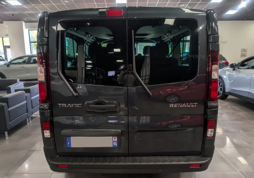 Vue arrière du Renault Trafic Combi gris comète, avec portes vitrées et essuie-glaces verticaux visibles en intérieur showroom.