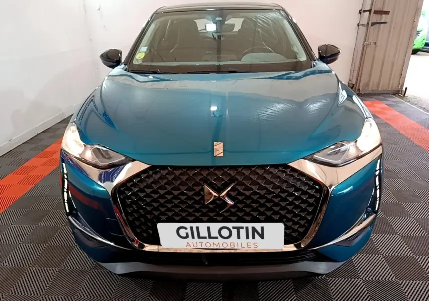 Vue frontale d'une DS3 Crossback bleu métallisé avec calandre noire à motifs géométriques et phares allumés.