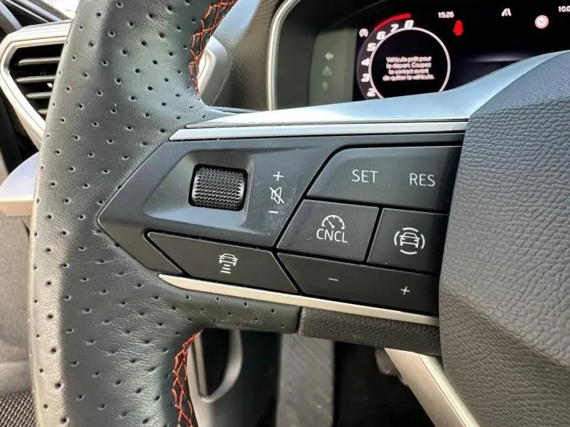 Gros plan sur les commandes au volant côté gauche du SEAT LEON ST 1.5 eTSI Hybrid 2025, cuir perforé noir avec surpiqûres rouges.