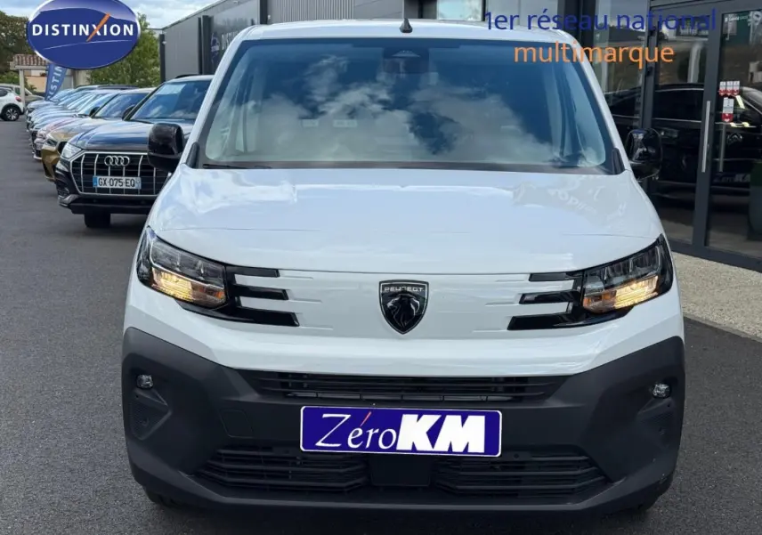 Vue frontale d'un Peugeot Partner blanc Icy 2026 avec feux allumés et pare-chocs noir mat visible en concession.