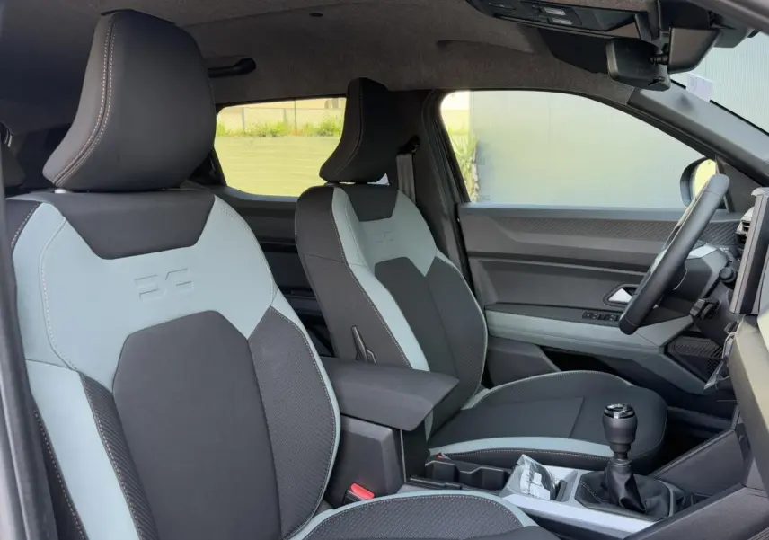 Intérieur avant droit du Dacia Duster 2025, sièges bi-ton gris clair et noir avec volant et levier de vitesse visibles.