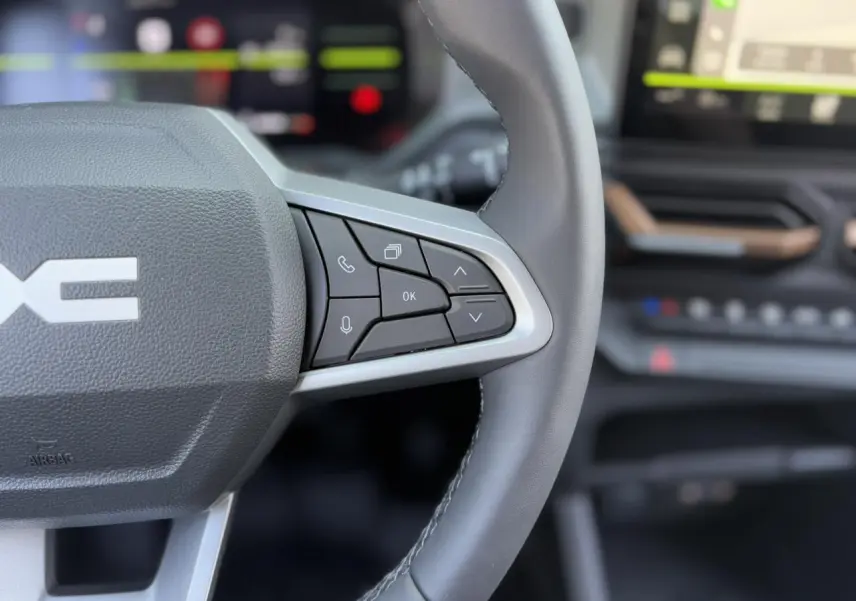 Gros plan sur le volant cuir noir du Dacia Duster 2025, avec commandes multifonctions et tableau de bord flou en arrière-plan.