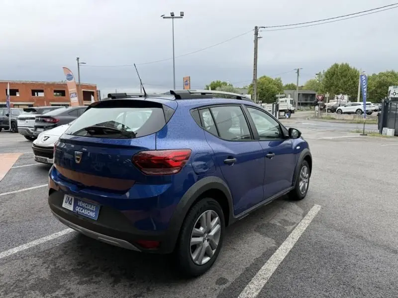 Vue 3/4 arrière droite d'une Dacia Sandero Stepway 2022 bleu métal avec barres de toit, garée en extérieur sur parking.