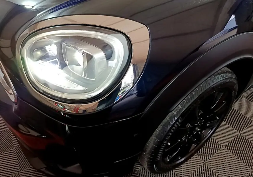 Gros plan sur le phare avant gauche et la roue noire d'une MINI Countryman Cooper SE bleu foncé.