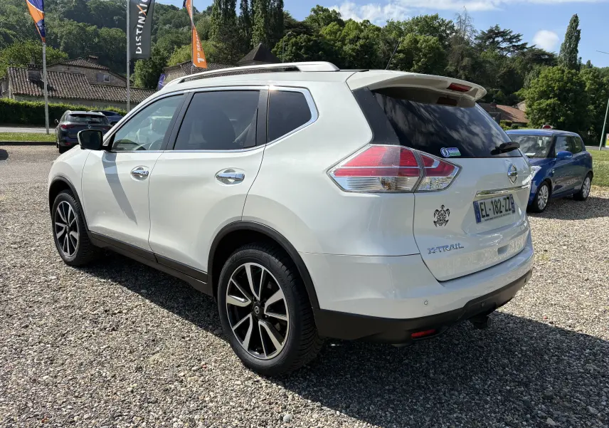 Nissan X-Trail blanc en 3/4 arrière droit, avec jantes noires et toit ouvrant, stationné sur un sol gravillonné.