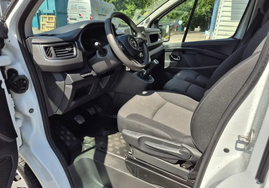 Vue intérieure côté conducteur du Nissan Primastar blanc, montrant le tableau de bord, volant multifonction et sièges en tissu gris.