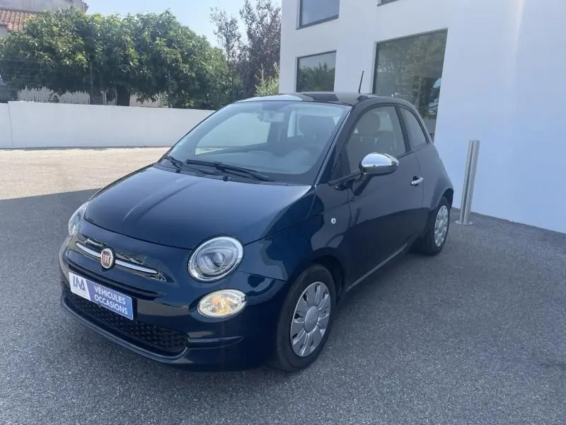 Fiat 500 bleu foncé métal vue 3/4 avant droit, avec phares allumés et rétroviseurs chromés, stationnée devant un bâtiment blanc.