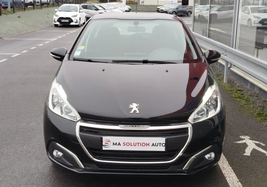 Vue de face d'une Peugeot 208 noire Perla Nera avec phares allumés et calandre chromée visible en extérieur.