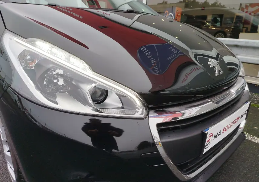 Gros plan sur l'avant gauche d'une Peugeot 208 noire Perla Nera, mettant en valeur le phare et le logo lion chromé.
