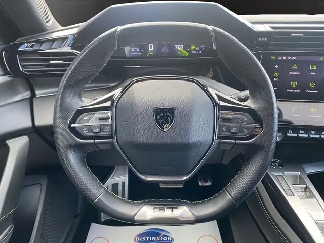 Vue rapprochée du volant cuir noir avec logo Peugeot et badge GT du poste de conduite de la 308 SW blanc nacré 2022.