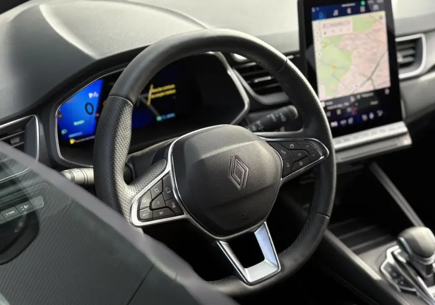 Vue intérieure centrée sur le volant noir perforé de la Renault SYMBIOZ 2025 avec écran tactile de navigation en arrière-plan.