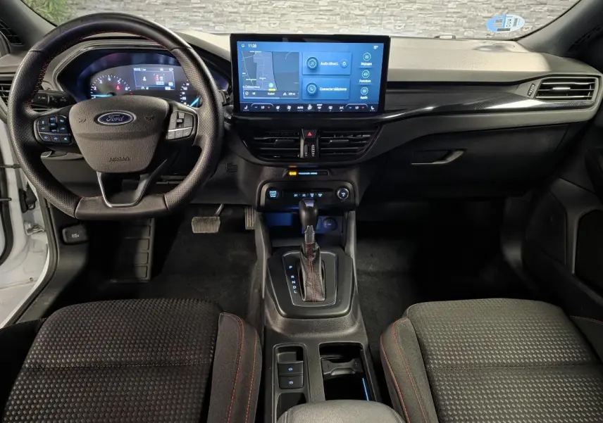 Vue intérieure avant de la Ford Focus 2024 blanche, tableau de bord noir avec écran tactile central et volant ST Line à surpiqûres rouges.