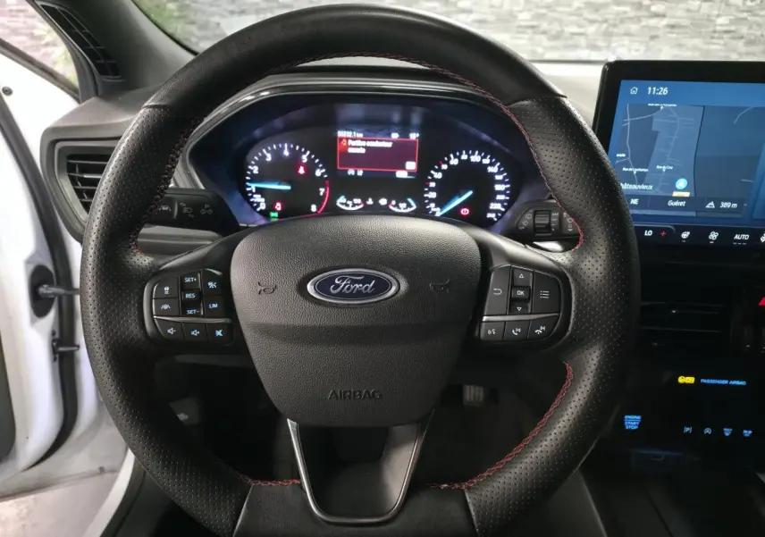 Vue intérieure centrée sur le volant perforé noir avec surpiqûres rouges et tableau de bord numérique d'une Ford Focus blanche 2024.