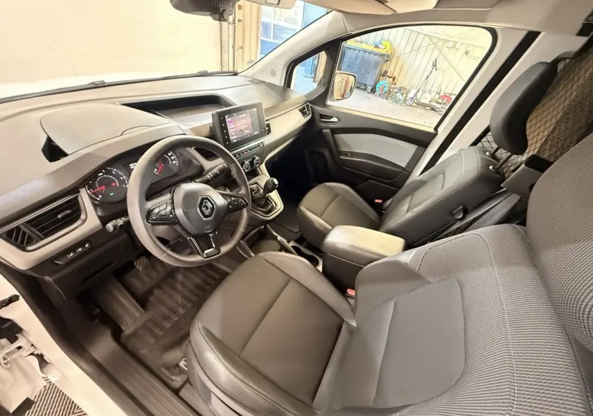 Vue intérieure côté conducteur du Renault Kangoo blanc 2022, montrant le tableau de bord et les sièges avant en tissu gris.