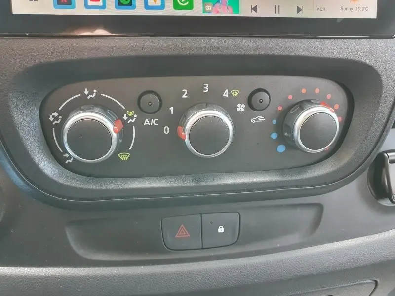 Gros plan sur les commandes de climatisation manuelle du Renault Trafic Fourgon blanc, avec boutons rotatifs noirs et indicateurs colorés.