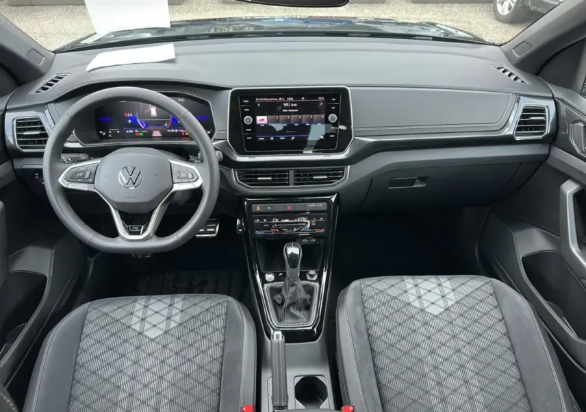 Vue intérieure avant du Volkswagen T-Cross TSI 116 DSG7 R-Line 2025, avec tableau de bord numérique et sellerie tissu grise à motifs.