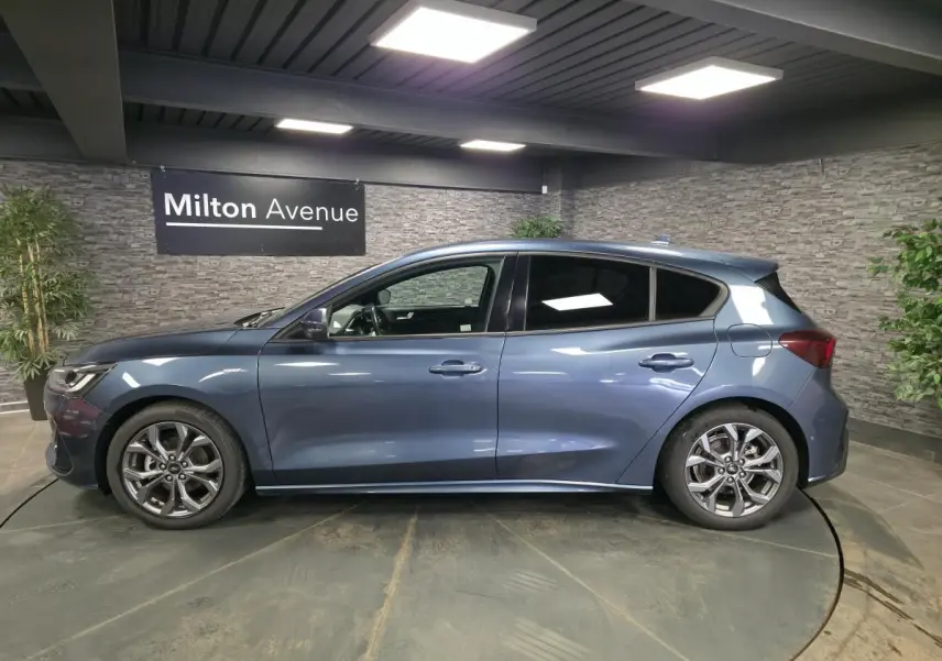 Profil gauche d'une Ford Focus 2024 bleu azur avec jantes alliage 17'' et vitres arrière surteintées dans un showroom.