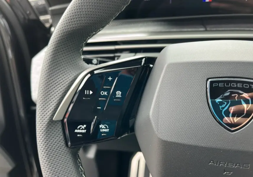 Gros plan sur le volant cuir perforé côté gauche du Peugeot 5008 Hybrid 145 GT avec logo lion et commandes intégrées.