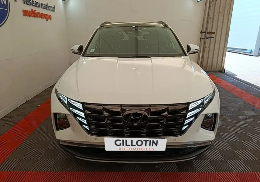 Vue frontale d’un Hyundai Tucson blanc 2022 avec calandre noire et feux LED distinctifs, en intérieur showroom.