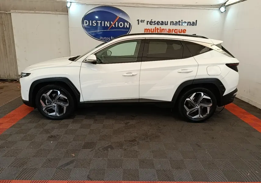 Vue latérale gauche d'un Hyundai Tucson blanc 2022 hybride, avec jantes alliage et toit noir contrasté en intérieur showroom.