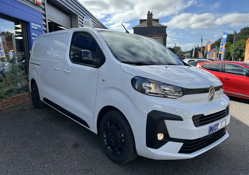 Vue 3/4 avant droit d’un Citroën Jumpy blanc avec jantes noires et calandre noire, stationné devant un bâtiment commercial.
