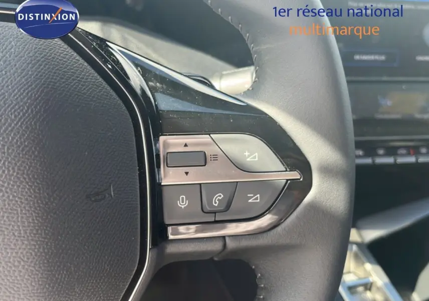 Gros plan sur les commandes au volant en cuir noir de la Peugeot 308 Hybrid 2025, avec tableau de bord flou en arrière-plan.