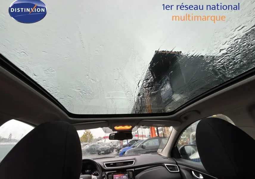 Vue intérieure avant du Nissan Qashqai blanc glacier 2017, avec toit panoramique et tableau de bord visible sous la pluie.