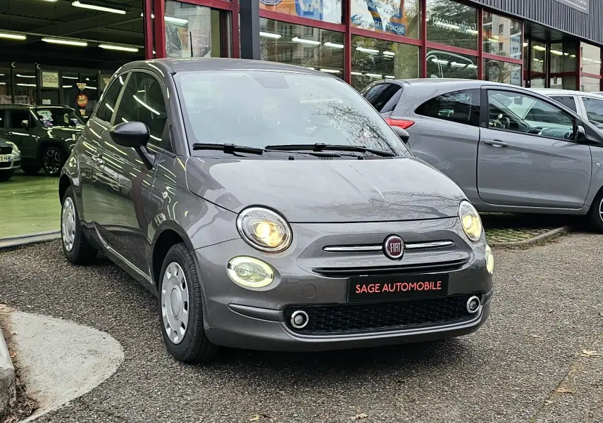 Fiat 500 gris vue 3/4 avant droit, phares allumés, stationnée devant un garage Sage Automobile.