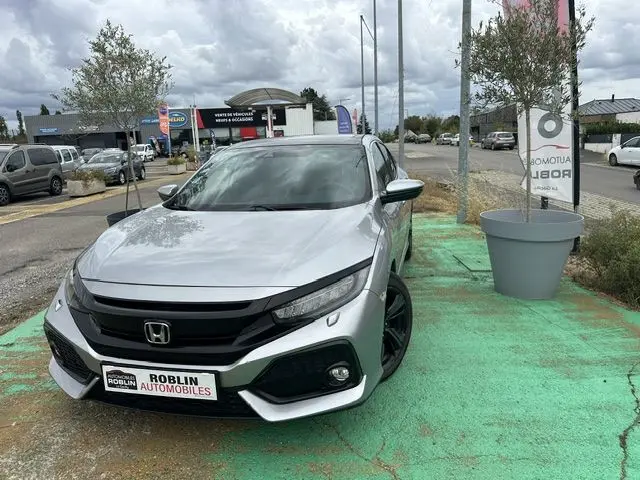 Vue 3/4 avant d'une Honda Civic grise stationnée, avec jantes noires et calandre noire distinctive.